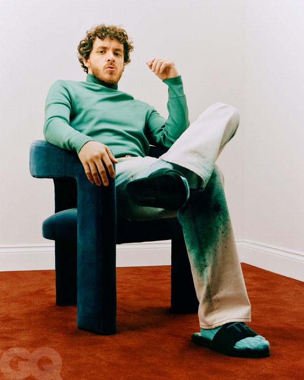 Rapper Jack Harlow sitting in a blue chair wearing a teal turtleneck, light denim jeans, and blue slides in a photo studio.