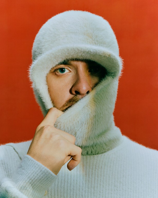 Close-up studio shot of an adult male wearing a light blue sweater and a furry hat with a bill against a red background.