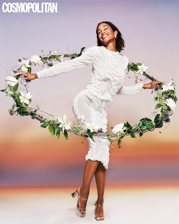 Smiling African-American woman in a white dress poses with a flower frame against a gradient background for a magazine cover.