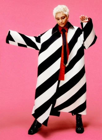 Young adult poses in a black and white diagonally striped overcoat and black patent boots against a pink backdrop in a fashion editorial.