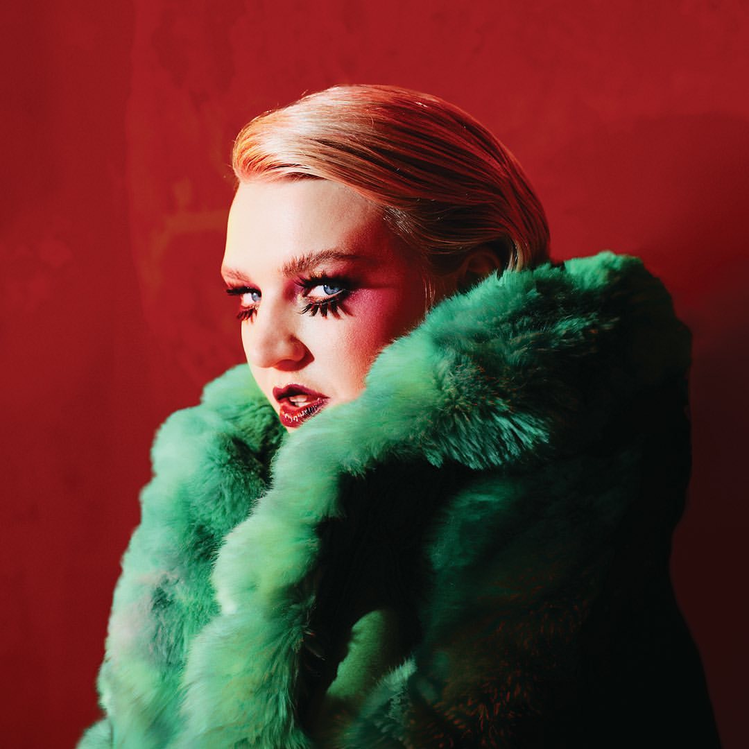 A woman with bright blue eyes and bold makeup, wearing a green fur coat against a vibrant red background, gazes intensely at the camera.