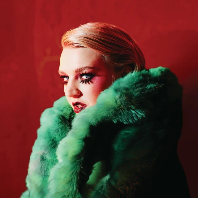 A woman with bright blue eyes and bold makeup, wearing a green fur coat against a vibrant red background, gazes intensely at the camera.