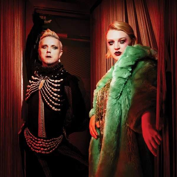 Two fashionably dressed people pose indoors with a red curtain backdrop, one in black skeleton attire and the other in a green faux-fur coat.