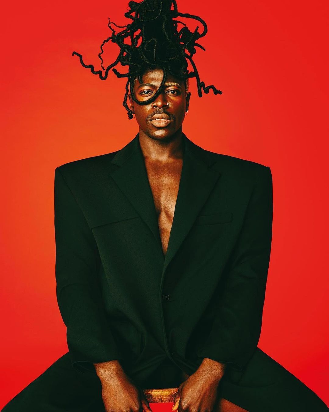 Portrait of an adult man with locs wearing a black blazer while seated against a vibrant red backdrop, captured in a medium shot.