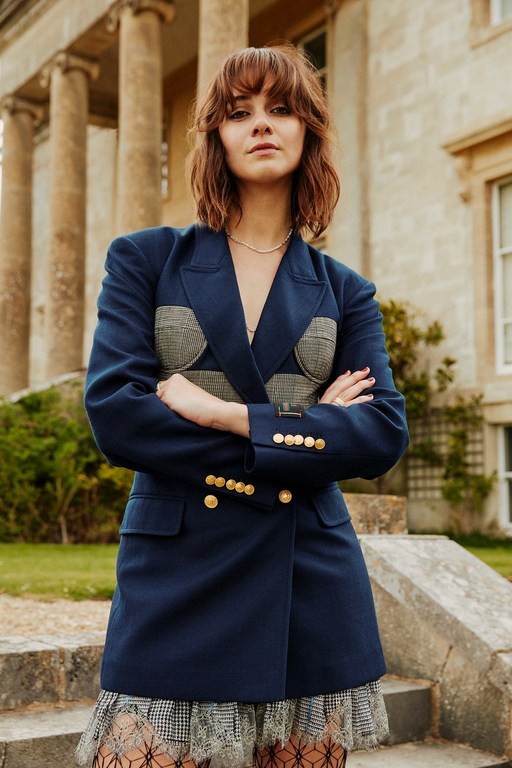 Portrait of a young woman with auburn hair wearing a navy blazer and short skirt, posing on stone steps in front of a building.
