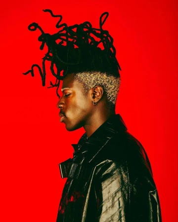 Profile portrait of a Black man with sculpted dreadlocks wearing a glossy black jacket against a bold solid red background.