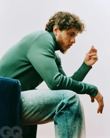 Jack Harlow seated in profile wearing a green turtleneck sweater and patterned denim jeans against a plain background
