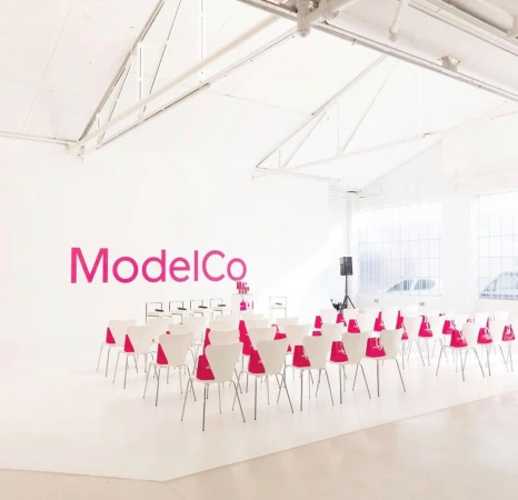 A photo studio with white walls, ample chairs, a speaker, and a large pink ModelCo logo, creating a bright and airy creative space.