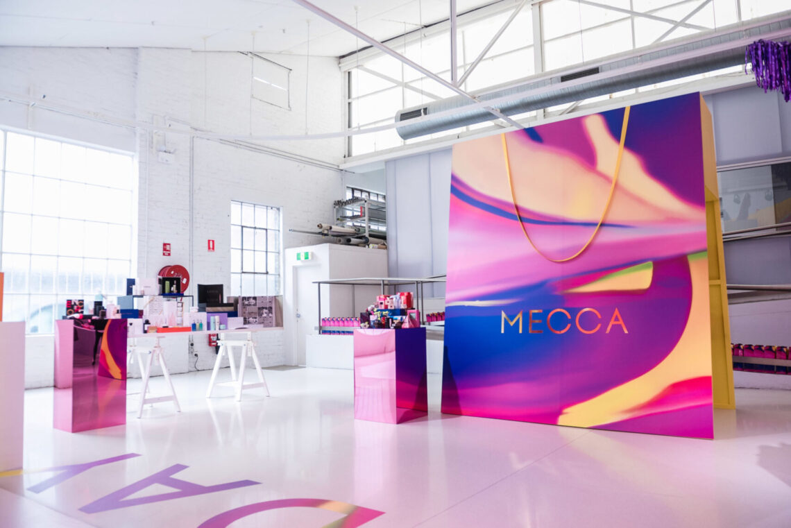 Interior view of a retail space with a large shopping bag prop, product display tables, and bright white floor. The 'MECCA' logo is visible.