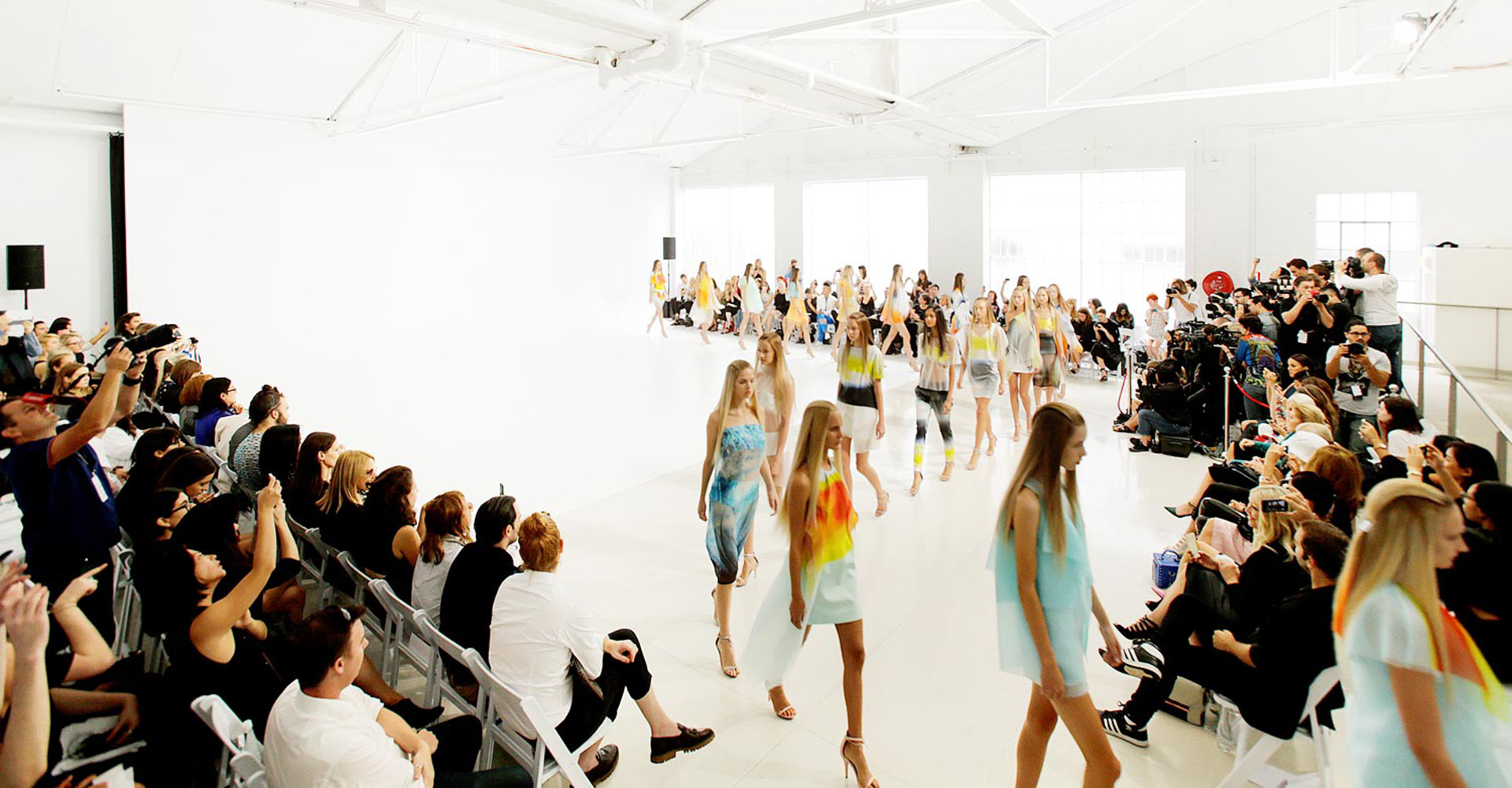 Fashion show event featuring models on the runway and audience members sitting on chairs in a brightly lit photo studio.