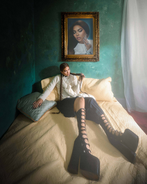 Woman with long braided hair wearing lace-up platform boots and white sweater seated on bed in green room.