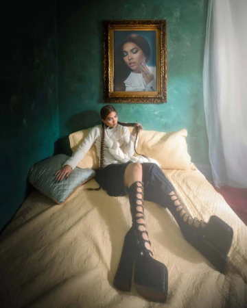 Woman with long braided hair wearing lace-up platform boots and white sweater seated on bed in green room.
