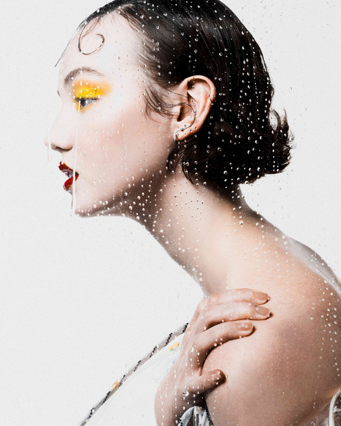 Profile portrait of a woman with yellow eyeshadow and red lips behind a glass surface covered in water droplets.