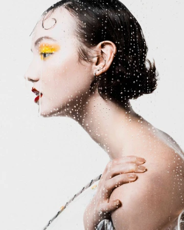 Profile portrait of a woman with yellow eyeshadow and red lips behind a glass surface covered in water droplets.