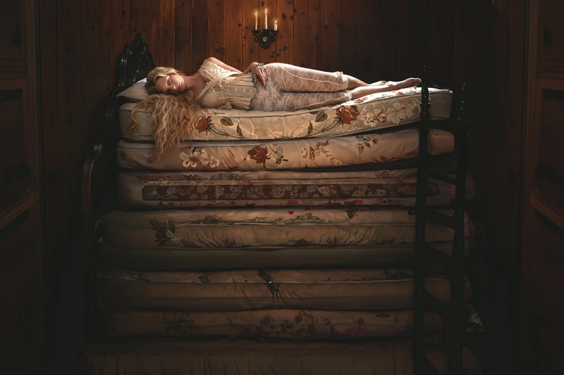 Woman in vintage attire rests atop a tall stack of floral mattresses in a dimly lit wood-paneled room with candlelight, creating a fairy-tale atmosphere.