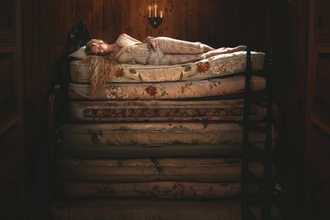 Woman in vintage attire rests atop a tall stack of floral mattresses in a dimly lit wood-paneled room with candlelight, creating a fairy-tale atmosphere.