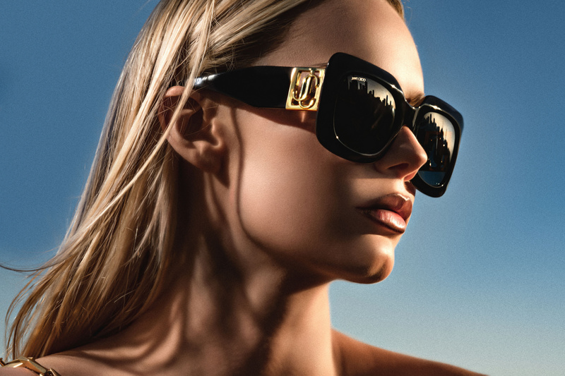 Close-up of a blonde woman wearing black sunglasses reflecting a cityscape against a clear blue sky, beauty shot, fashion photography.