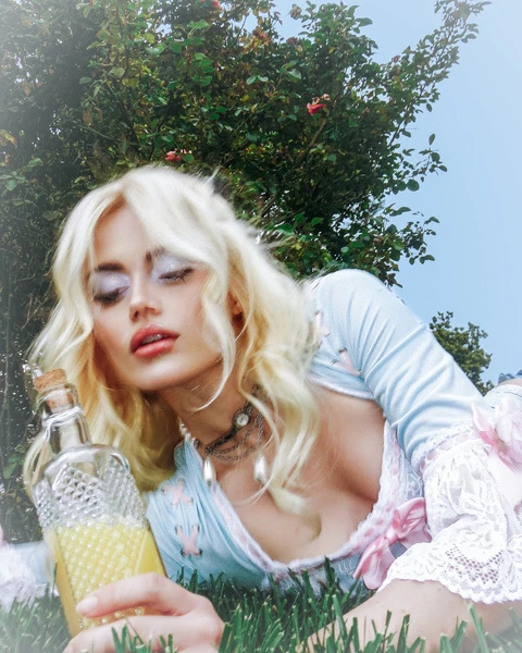 Woman with blond hair laying on grass, holding a bottle with yellow liquid, wearing a light blue top in an outdoor setting.