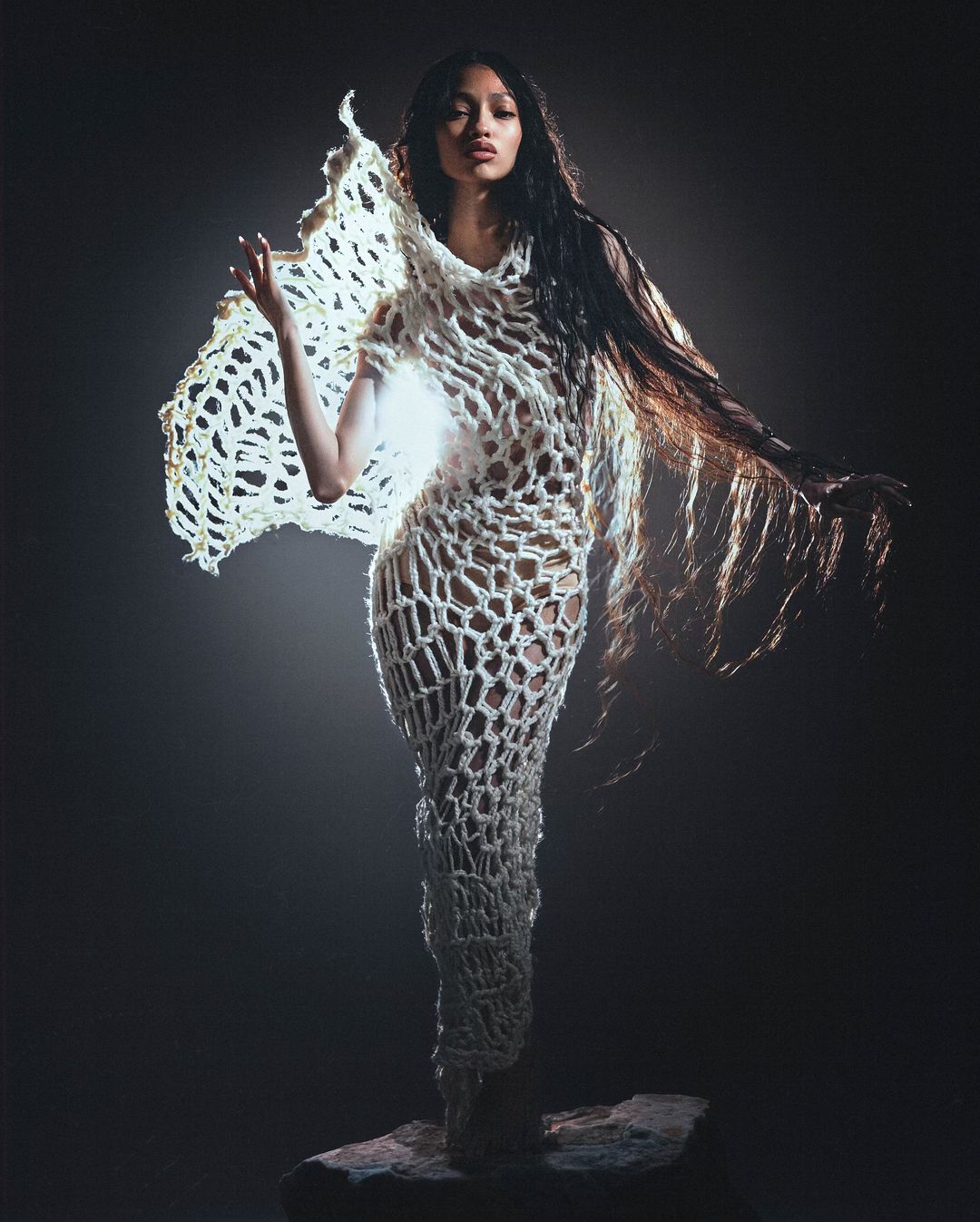 A woman with long dark hair stands on a stone wearing a white crochet dress against a dark background in a fashion photography image.