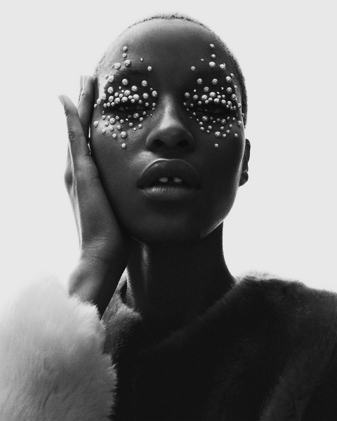 Monochrome portrait of a Black woman with pearl embellishments around her eyes, touching her cheek.