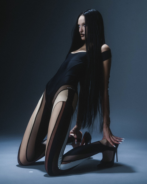 Model in black bodysuit and sheer leggings poses in studio with dramatic lighting and long dark hair for a fashion editorial shoot.