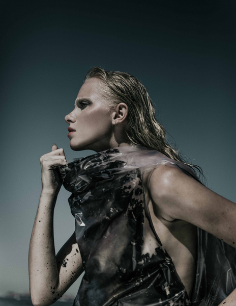 Adult woman with wet blonde hair and dramatic makeup poses in translucent plastic fashion in a dramatic side-lit outdoor fashion shot.
