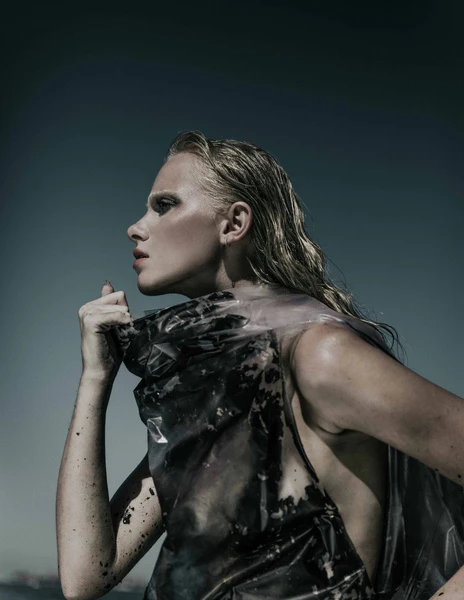 Adult woman with wet blonde hair and dramatic makeup poses in translucent plastic fashion in a dramatic side-lit outdoor fashion shot.
