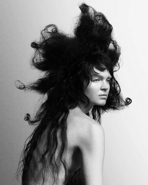 Striking black and white portrait of a bare-shouldered individual with an elaborate, dark, voluminous hairstyle against a neutral backdrop.