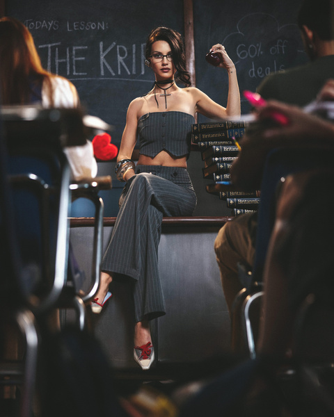 Female fashion model poses in a classroom setting with a blackboard background and fellow students, wearing gray pinstripe outfit.