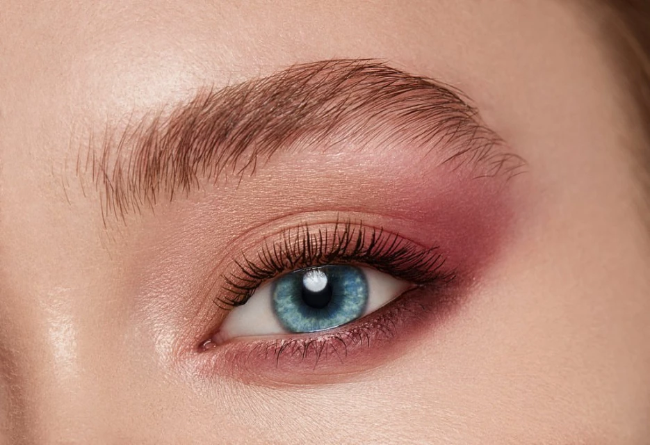 Close-up studio shot of a woman's face featuring an eye with blue iris, long eyelashes, a styled eyebrow, and pink eyeshadow.