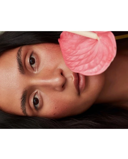 Close-up portrait of an adult woman with dark hair and brown eyes, featuring a pink anthurium flower in the foreground.