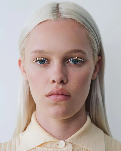 Close-up portrait of a fair-skinned woman with platinum blonde hair, light-colored clothing, and rhinestone decorations under her eyebrows.