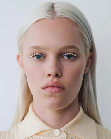 Close-up portrait of a fair-skinned woman with platinum blonde hair, light-colored clothing, and rhinestone decorations under her eyebrows.