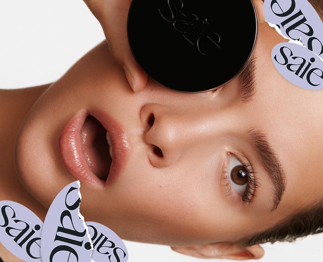 Close-up studio shot of a fair-skinned adult female with brown eyes and an open mouth holding a black cosmetic product container.