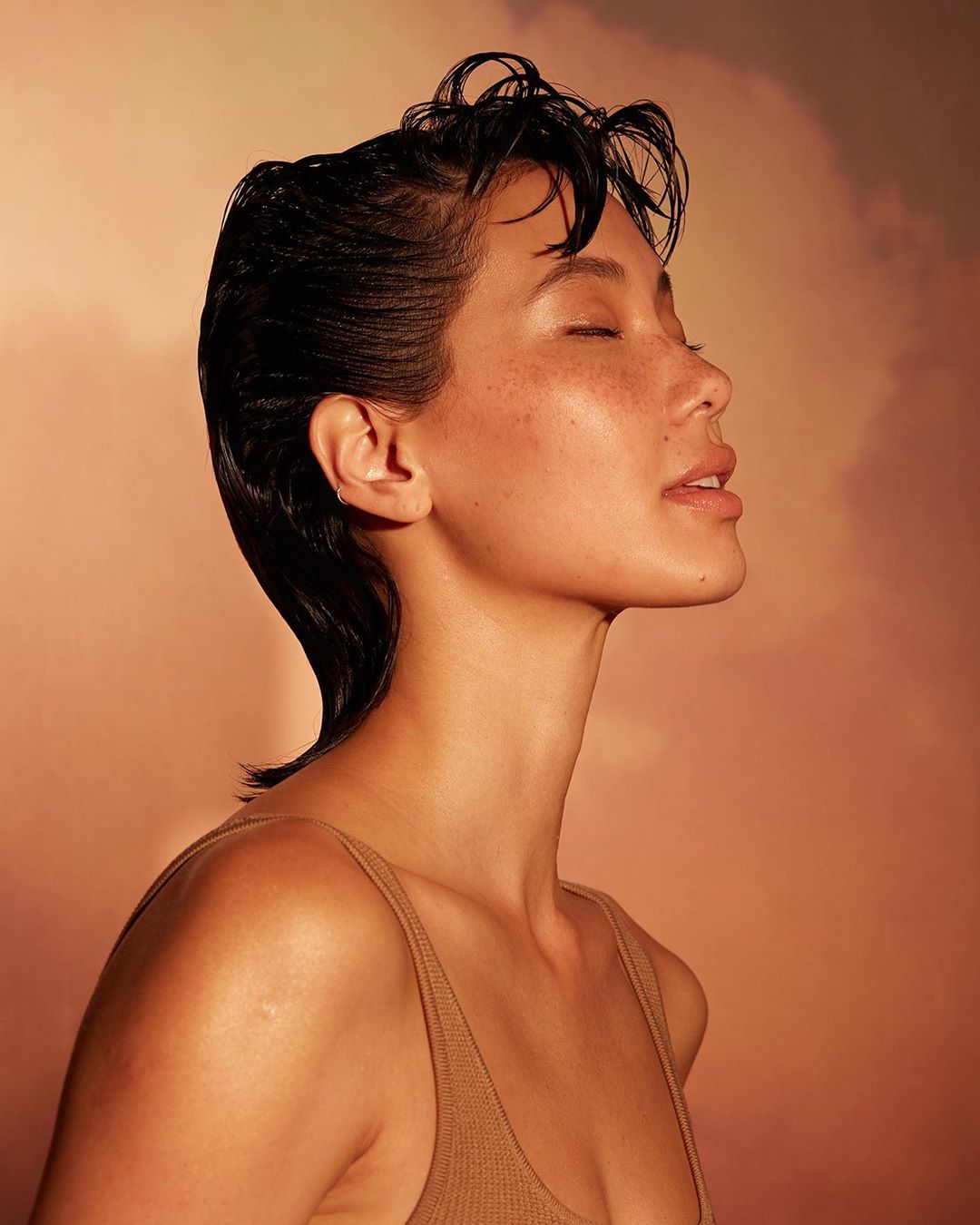 Portrait of a young woman with short, dark, wet hair, eyes closed, bathed in warm, soft lighting against a peach-toned background.
