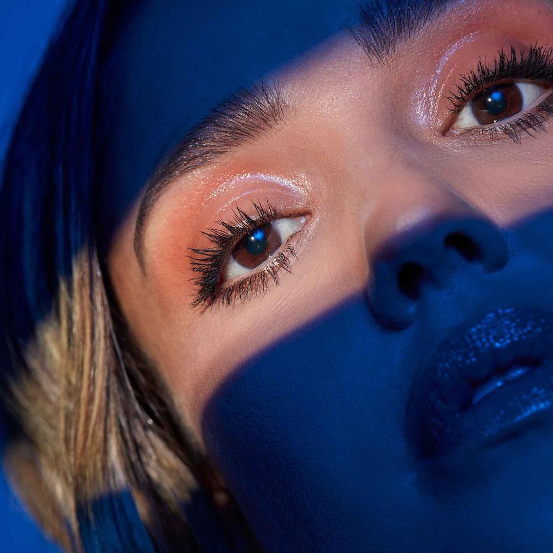 Close-up shot of an adult woman's face with brown eyes, flawless makeup, glossy lips, and a blue-toned light casting a shadow.