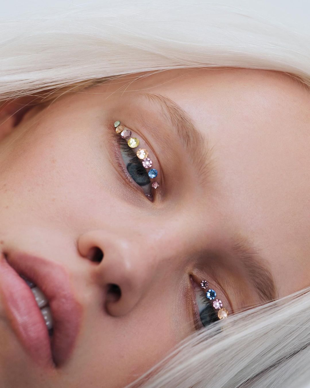 Close-up of a light-skinned person with blue eyes and white hair, adorned with colorful rhinestones around the eyes in a beauty shot.