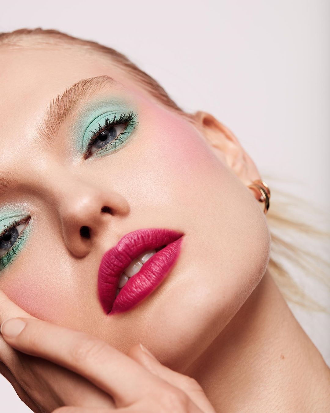 Close-up portrait of a young fair-skinned woman with mint-green eyeshadow, pink blush and dark pink lips in a bright, neutral setting.
