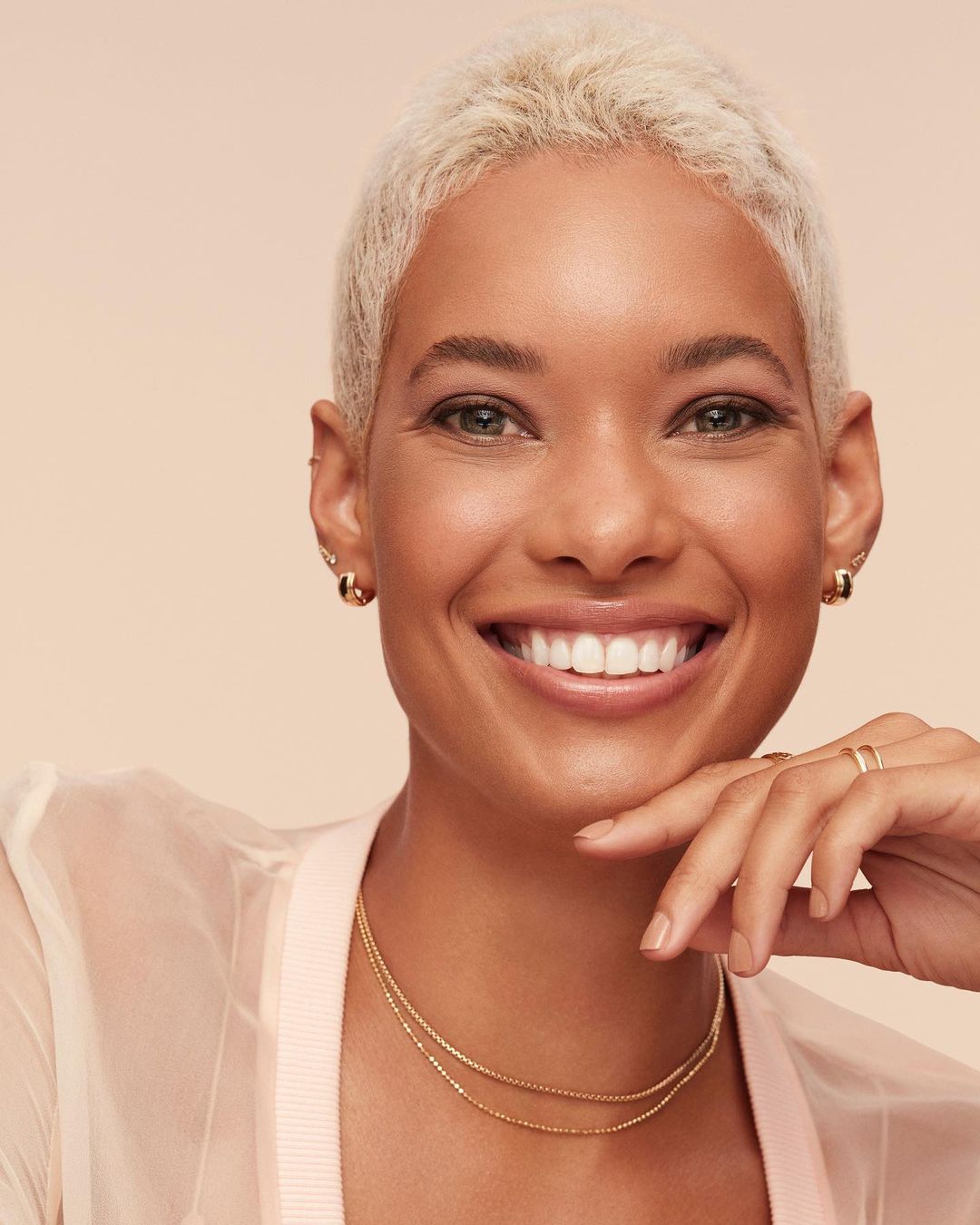 Close-up shot of a smiling adult woman with short blonde hair, wearing gold jewelry and a sheer peach-colored jacket, posing indoors.