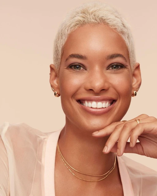 Close-up shot of a smiling adult woman with short blonde hair, wearing gold jewelry and a sheer peach-colored jacket, posing indoors.