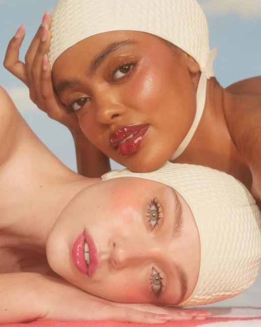 Close-up of two women with glossy makeup and vintage swim caps against a soft sky, showcasing retro beauty and skincare.