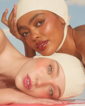Close-up of two women with glossy makeup and vintage swim caps against a soft sky, showcasing retro beauty and skincare.