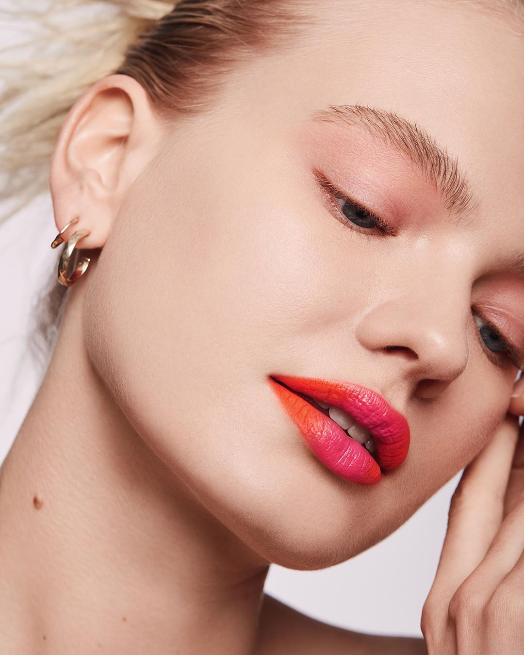 Close-up portrait of a fair-skinned woman with orange and pink ombre lips and gold hoop earrings against a white background.