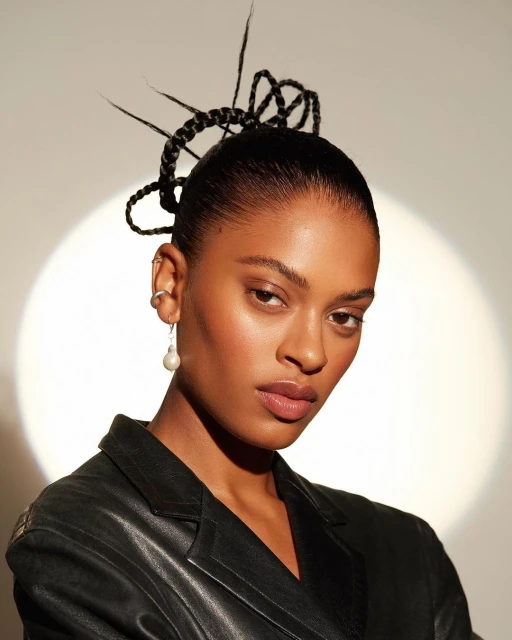 Close-up portrait of a young woman with braided hair wearing a black jacket against a bright background. Pearl drop earring visible.
