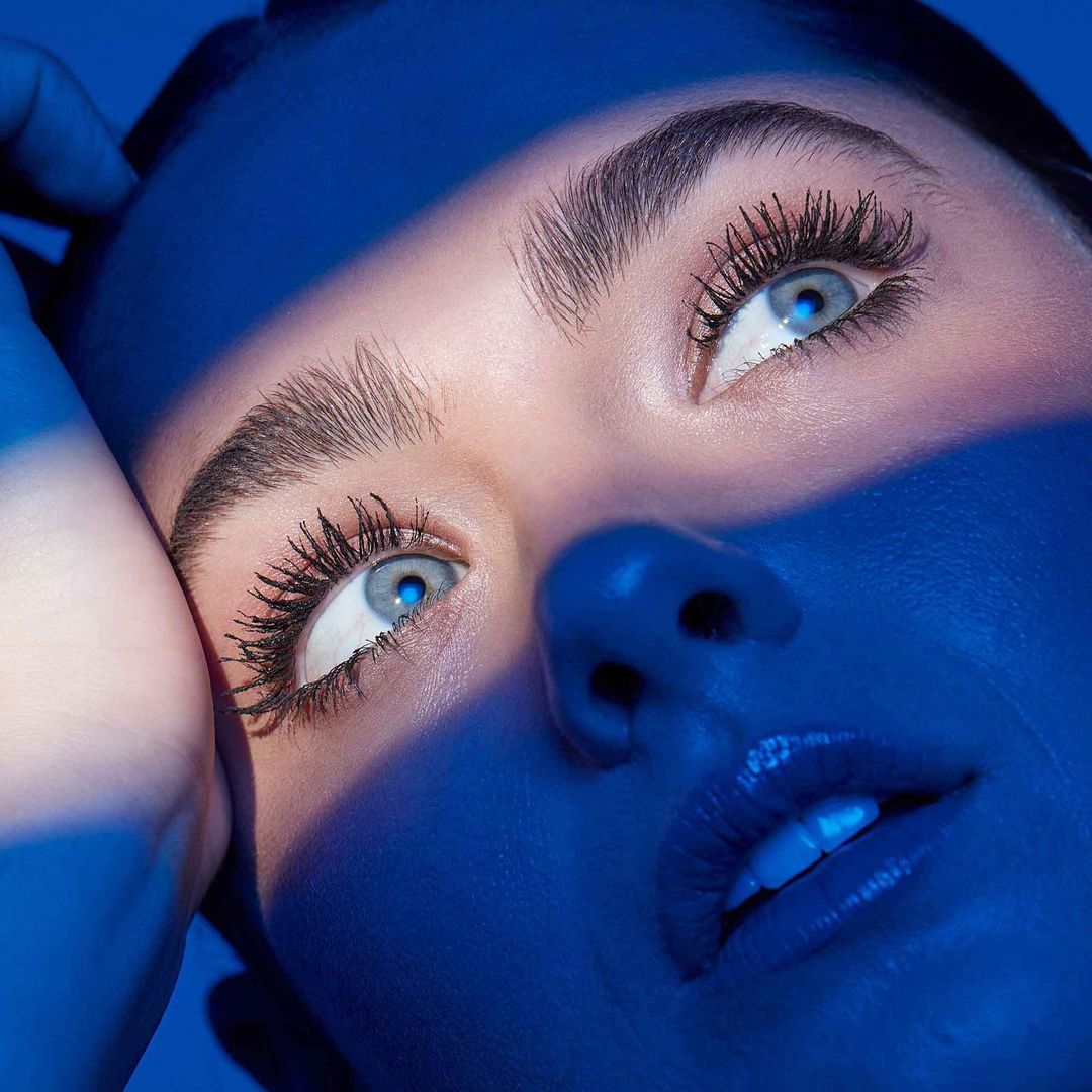 Close-up of a woman's face in blue lighting showcasing her eyes, eyebrows, eyelashes, and glossy lips against a solid blue backdrop.