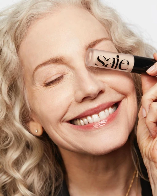 Close-up of an elderly woman smiling, holding a Saie makeup product near her eye in a bright, clean studio environment. Beauty and cosmetics focus.