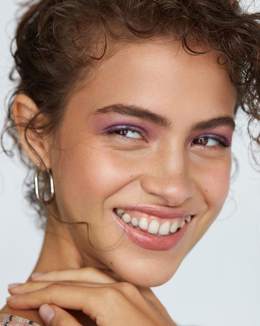 Close-up of a smiling woman with curly hair and purple eyeshadow. The focus is on her joyful expression and beauty makeup.