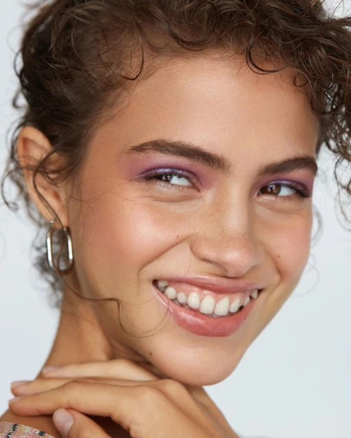 Close-up of a smiling woman with curly hair and purple eyeshadow. The focus is on her joyful expression and beauty makeup.