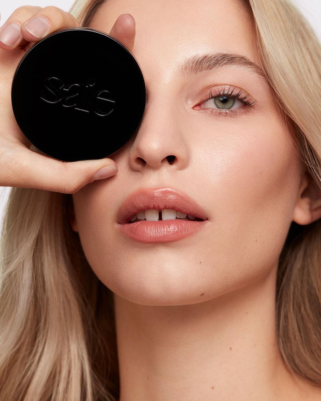 Close-up of a blonde woman holding a black cosmetic container over her eye, showcasing beauty and skincare in a bright studio setting.