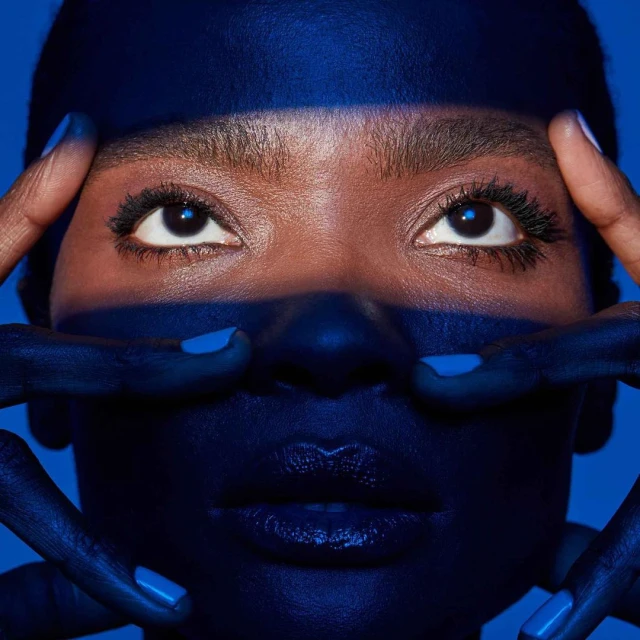 Close-up portrait of a Black woman with blue skin and blue lipstick, framing her face with hands painted in blue, against a blue background.
